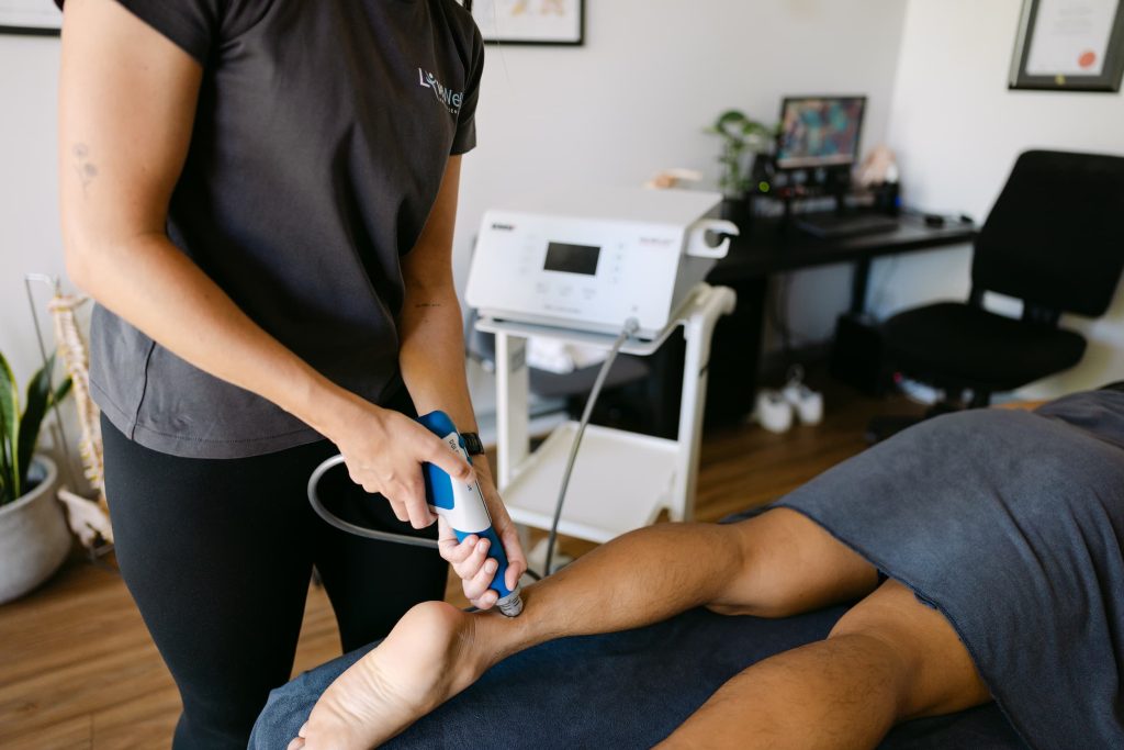 Osteopath performing shockwave therapy on Achilles tendon in clinic treatment room.