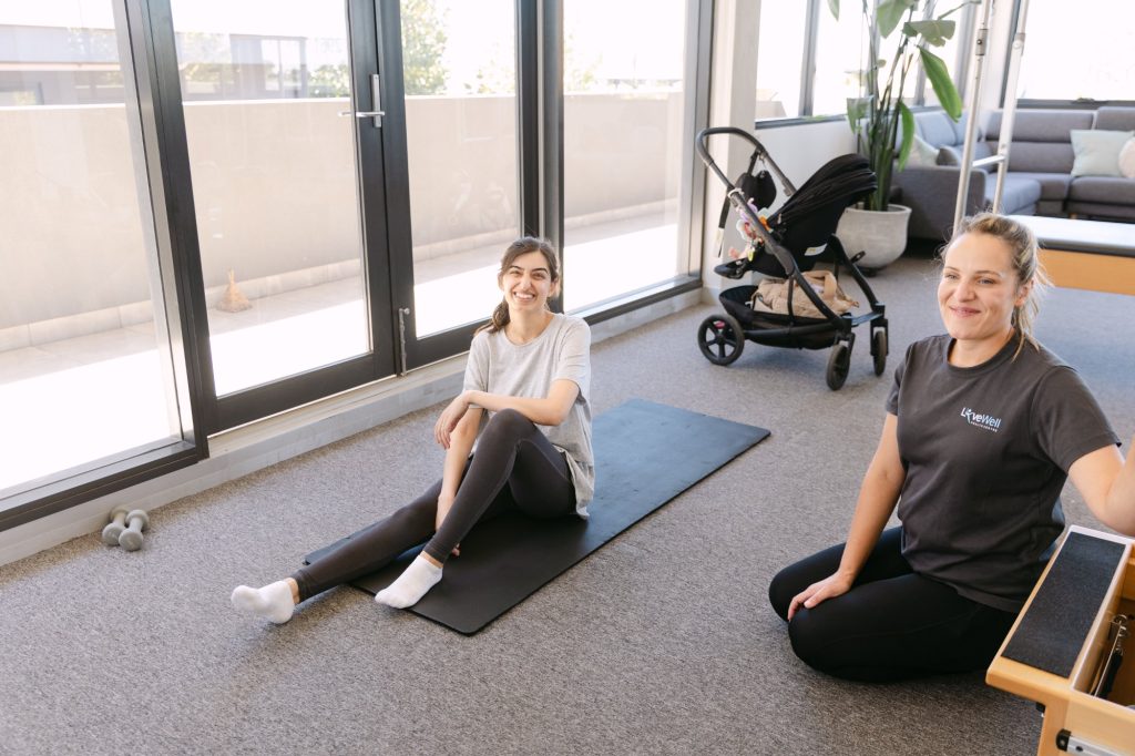 Live Well practitioner and postnatal patient smiling during mat Pilates session with pram nearby
