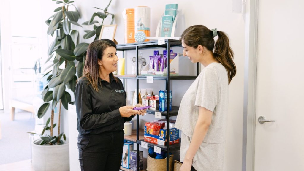Live Well staff member assisting postnatal patient with health products in bright clinic corridor